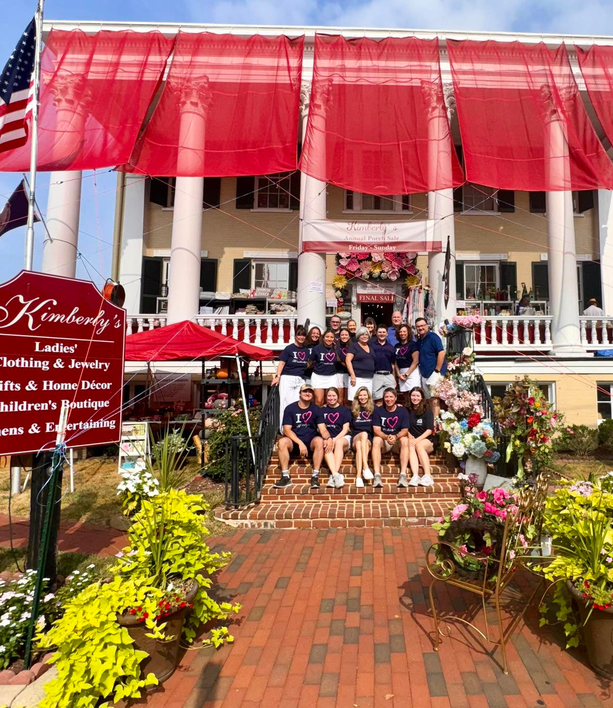 Destination shopping and dining in Winchester, VA. Photo shows Kimberly's Boutique staff in matching navy "I heart" t-shirts, posing on the steps of the boutique's historic building. It is set up for sale with large red awnings shading the porch.
