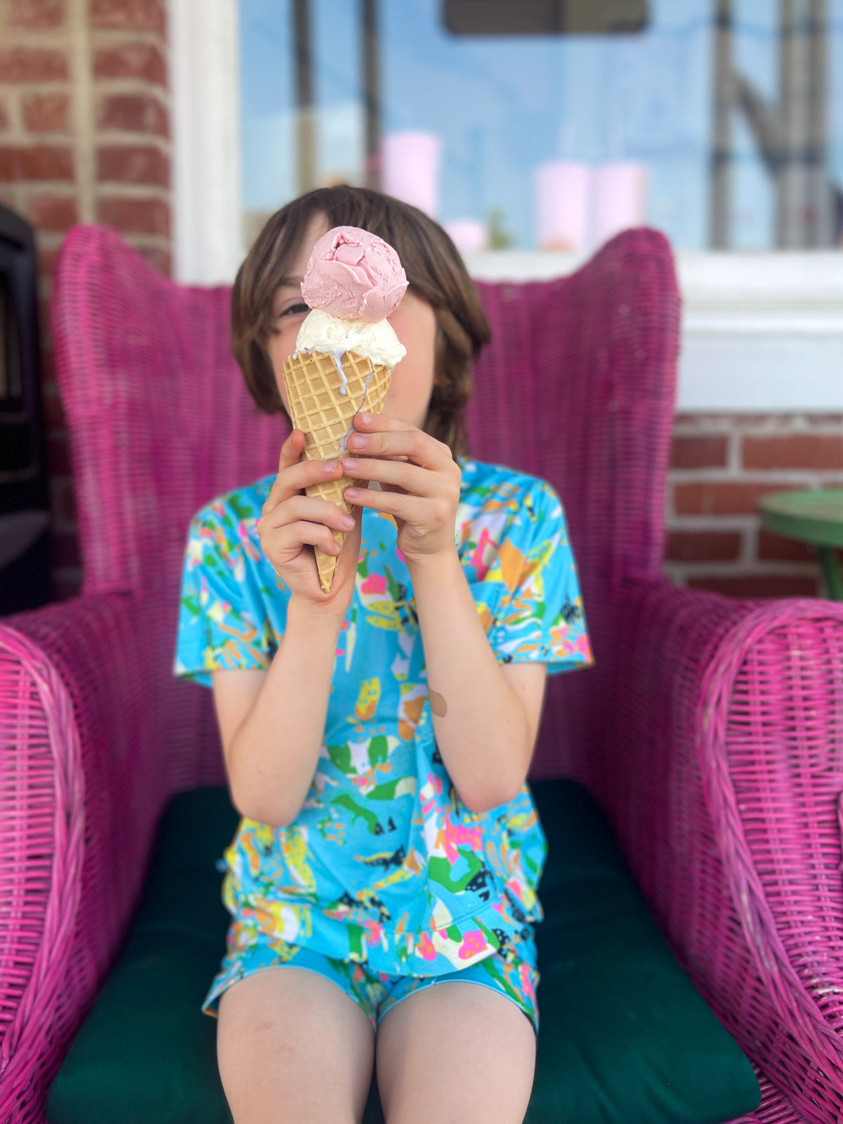 A child wearing a bright blue tropical print short-sleeve shirt and matching shorts sits in a pink wicker chair, holding a pink ice cream cone in front of their face.