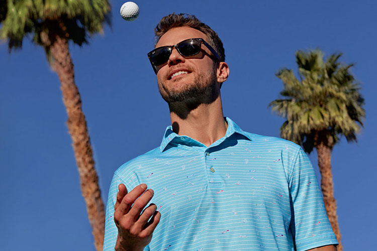 Man in sunglasses and blue shirt with a golf ball against a clear blue sky with palm trees.