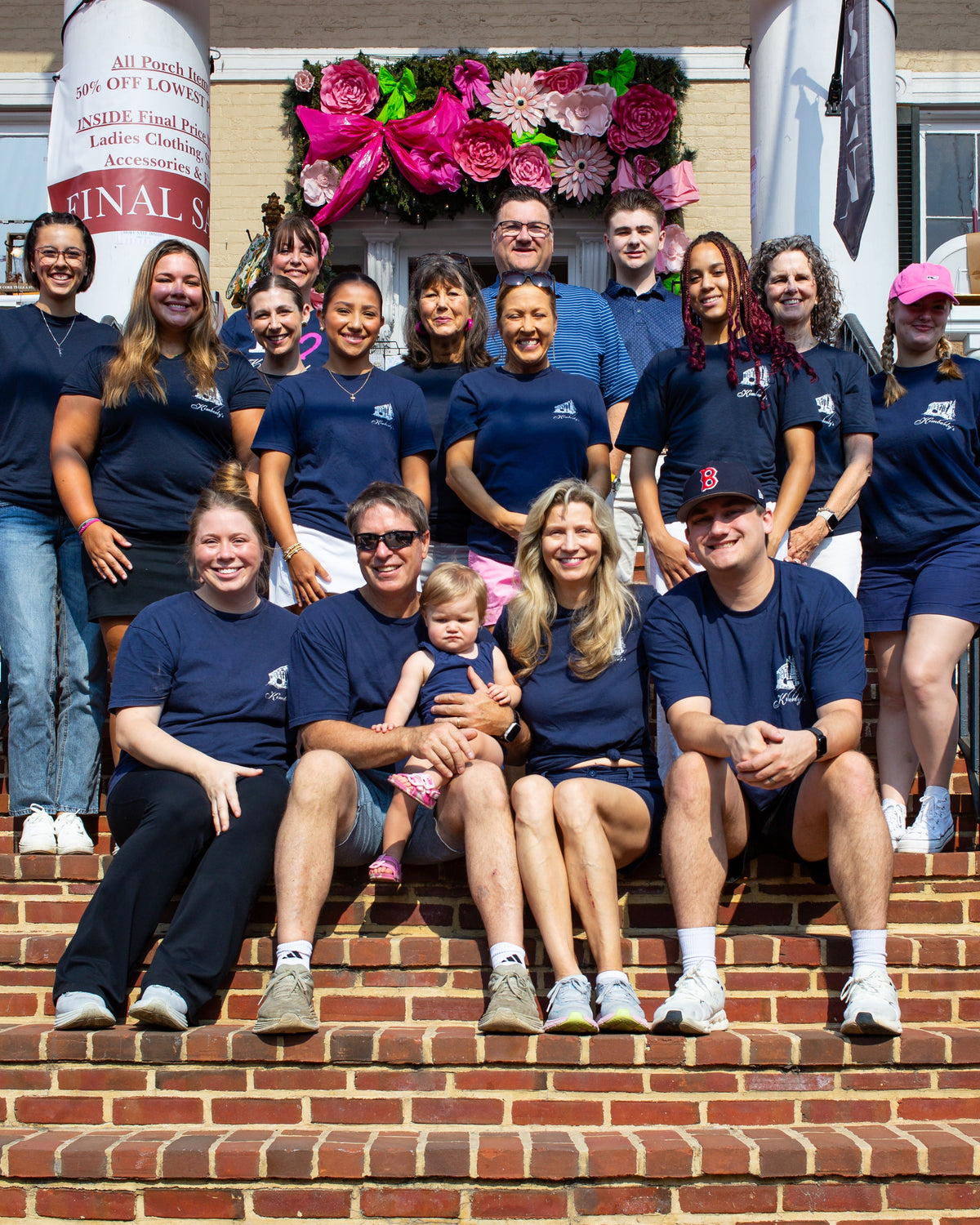 A group of people posing together on the brick steps of a building, wearing matching navy blue t-shirts with a Kimberly's logo. The background shows the Kimberly's boutique building facade with a balcony, black railings, and a decorative floral arrangement.