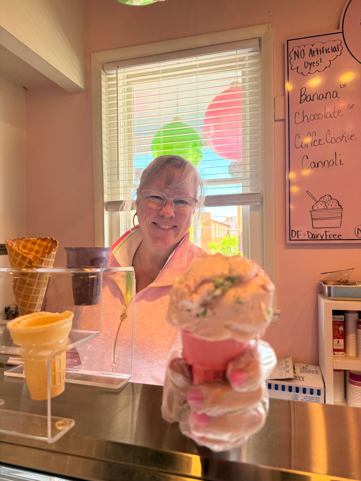 A smiling woman in a pink shirt stands behind a counter in a pastel pink ice cream shop, holding a large ice cream cone in front of her. Waffle cones and a chalkboard menu are visible in the background.