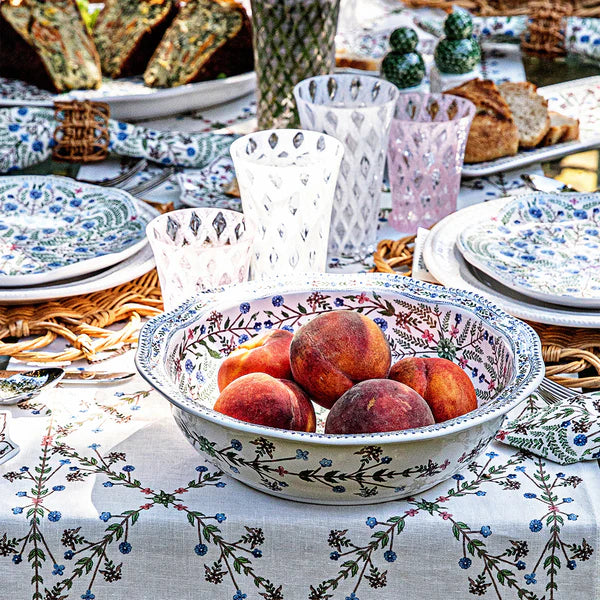 a full table setting closeup with dainty matching floral tablecloth and serving bowls on a white background. Colorful white and pink blown glass lattice tumblers sit behind the serving bowl.